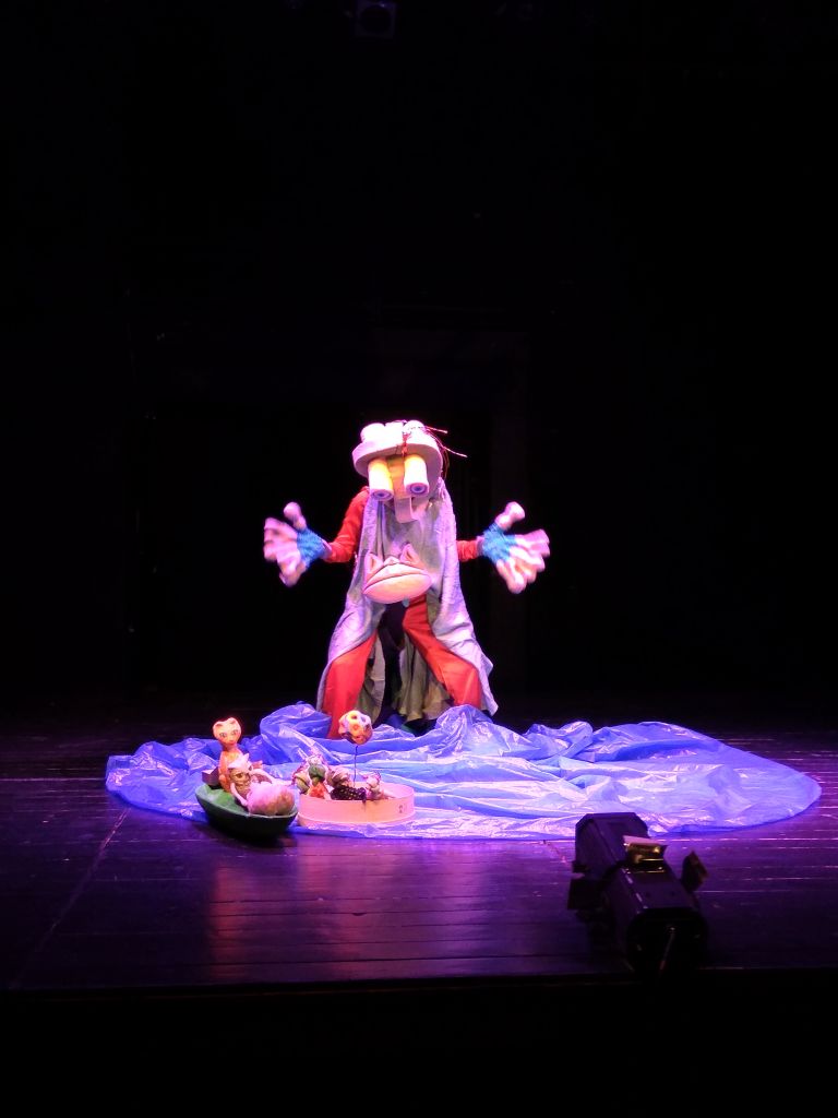 Στο κέντρο της σκηνής μια φιγούρα με τεράστιες παλάμες και μία μάσκα με μεγάλα διογκωμένα μάτια σαν κυλίνδρους στο κεφάλι έχει στην κοιλιά της ένα κεφάλι βατράχου και στέκεται με τα πόδια και τα χέρια ανοιγμένα πάνω από ένα κυματιστό γαλάζιο πανί όπου φαίνονται να πλέουν δύο μικρές βαρκούλες.