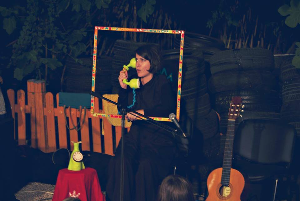 Μπροστά από μία στοίβα λάστιχα αυτοκινήτων και έναν ξύλινο χαμηλό φράχτη είναι καθισμένη μια κοπέλα με μαύρα ρούχα που μιλά σε ένα παράξενο κίτρινο τηλέφωνο. Γύρω από το πρόσωπο και το μπούστο της στηρίζει μια πολύχρωμη κορνίζα που την πλαισιώνει.