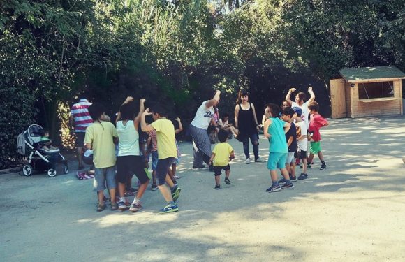 Στο απογευματινό φως του κατάφυτου θεάτρου της Ρεματιάς δύο ομάδες παιδιών χοροπηδούν ξέφρενα δίπλα σε δύο γυναίκες που στέκονται απέναντί τους. Στο βάθος δεξιά υπάρχει ένα μικρό ξύλινο κιόσκι με πράσινη σκεπούλα.