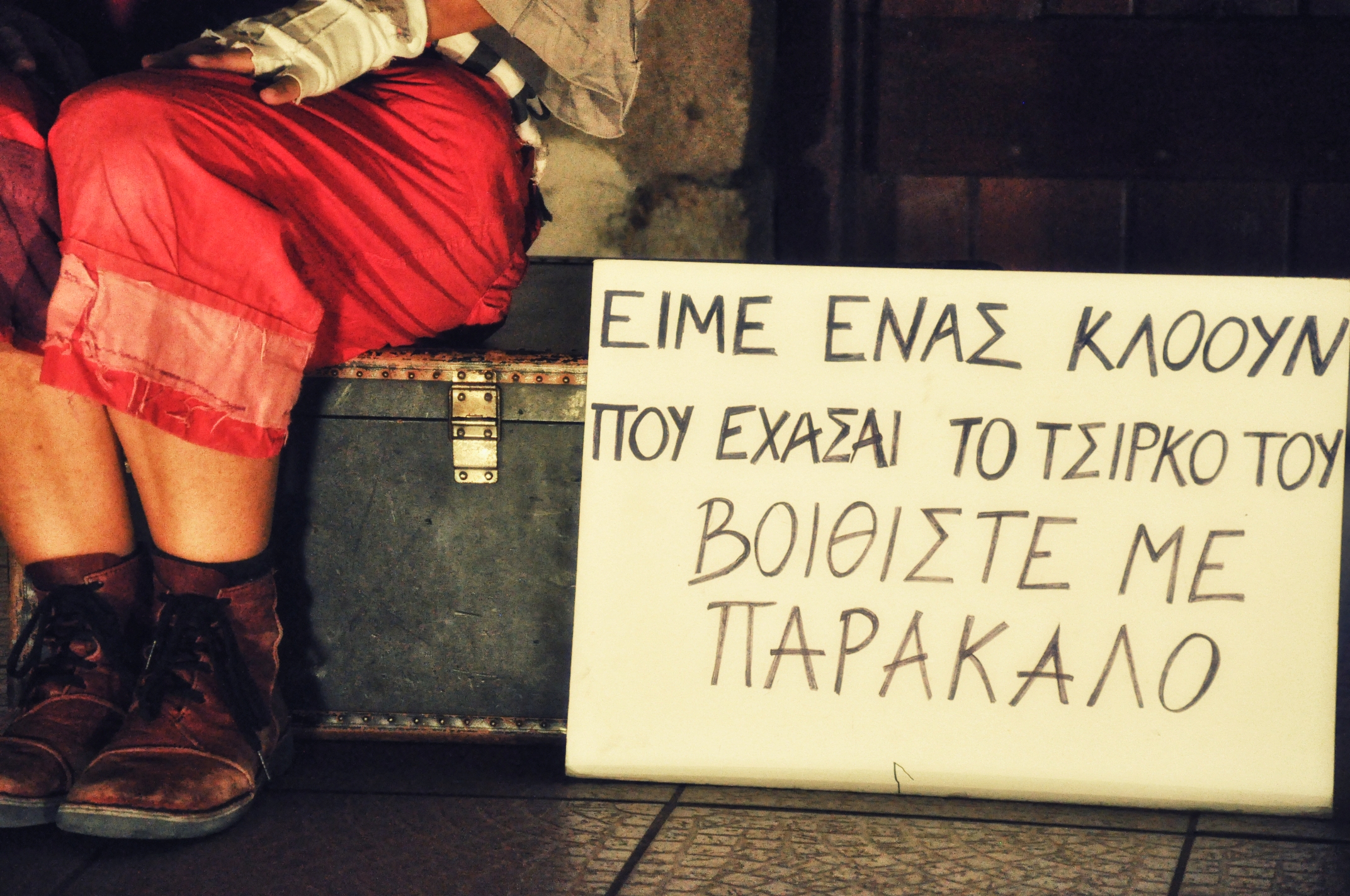 Πάνω σε μία παλιά πράσινη βαλίτσα είναι καθισμένος ένας άνθρωπος του οποίο βλέπουμε μόνο από τη μέση και κάτω. Φορά φθαρμένα μπορντώ μποτάκια και ένα κάπρι κοκκινωπό παντελονάκι με μπαλώματα. Δίπλα του στηριγμένο στη βαλίτσα είναι ένα πλακάτ όπου είναι ανορθόγραφα γραμμένο: Είμαι ένας κλόουν που έχασε το τσίρκο του βοηθήστε με παρακαλώ.