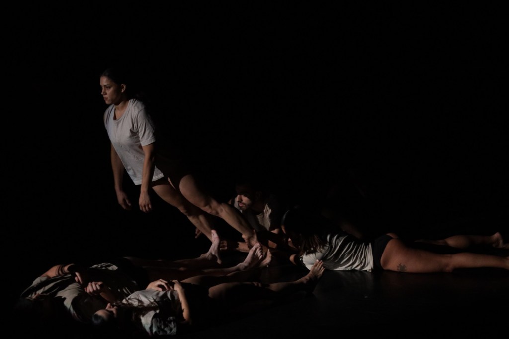 Δύο χορευτές ξαπλωμένοι ανάσκελα και δύο άλλοι ξαπλωμένοι μπρούμυτα συγκρατούν από τους αστραγάλους το σώμα μίας μελαχρινής χορεύτριας που γερμένη σα σανίδα προς τα μπρος έχει βγει τελείως εκτός άξονα.