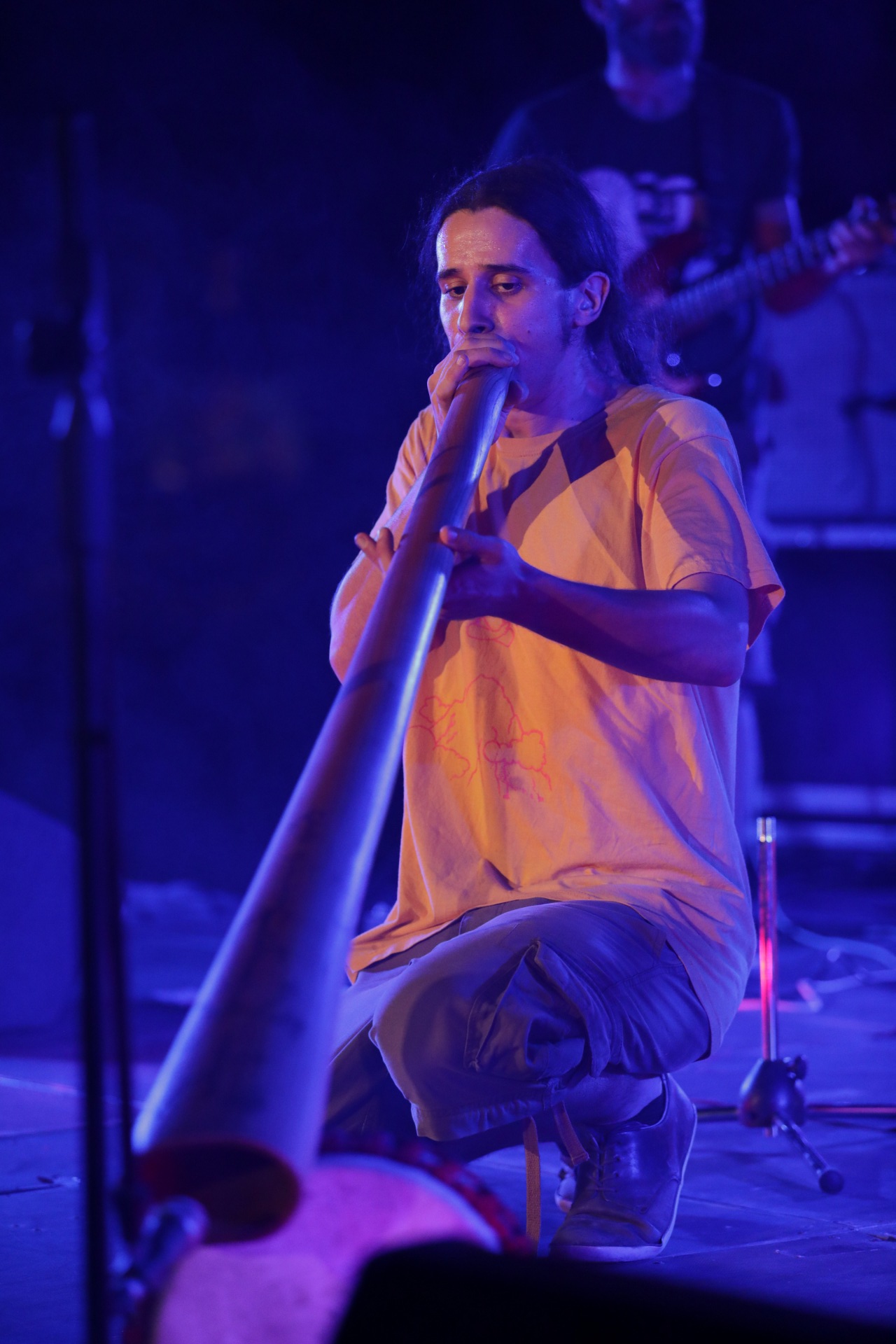 Κάτω από τον μπλε απαλό φωτισμό της μουσικής σκηνής, ένας άντρας με κοτσίδα και καλοκαιρινό φαρδύ t-shirt έχει γονατίσει προς το έδαφος και φυσά ένα μεγάλο didjeridoo, ενώ στο βάθος πίσω του ένας άλλος άντρας παίζει ηλεκτρική κιθάρα.