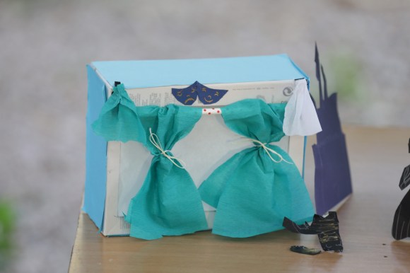 Ένα χάρτινο κουτί παπουτσιών έχει στολιστεί με βεραμάν κουρτινάκια από γκοφρέ χαρτόνι και δύο μικρές μπλε σκούρες μάσκες θεάτρου από κανσόν και έχει μετατραπεί σε θεατρική σκηνούλα.