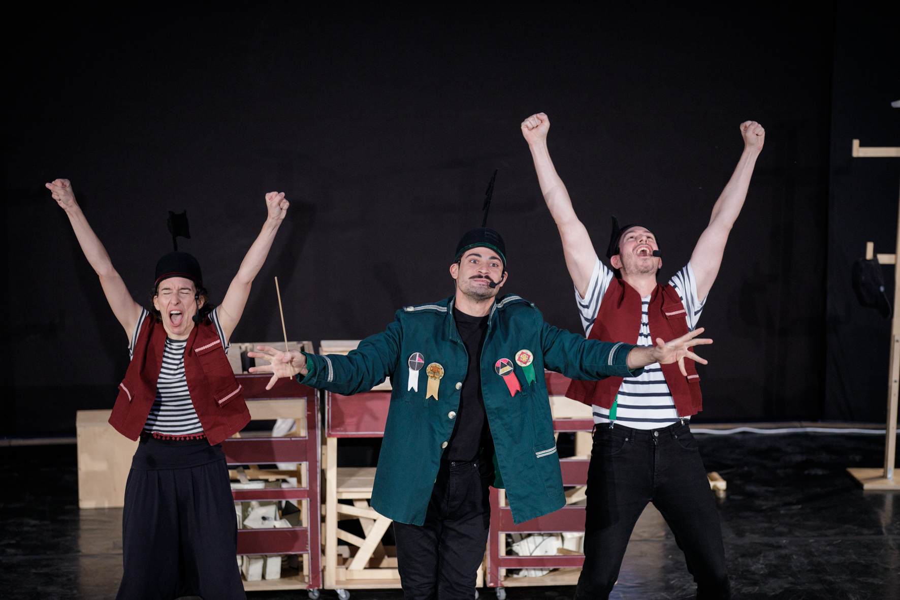 Στη σκηνή του θεάτρου της Ρεματιάς, μία κοπέλα και ένα αγόρι με μπορντώ γιλέκα και μαύρα στρογγυλά καπέλα σηκώνουν θριαμβευτικά τις γροθιές τους στον αέρα με το στόμα τους ορθάνοιχτο. Ανάμεσά τους, ένας νεαρός άντρας με κυπαρισσί σακάκι γεμάτο παράσημα και παρόμοιο καπέλο έχει τα χέρια ανοιγμένα στο πλάι σα μαέστρος μπροστά από την ορχήστρα.