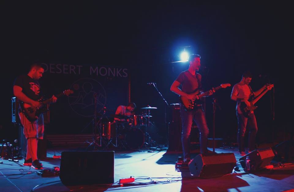 Αριστερά ένας άντρας παίζει κιθάρα, στη μέση ένας ακόμα παίζει κιθάρα και τραγουδάει, δεξιά του ένας τρίτος παίζει μπάσο και πίσω τους το τέταρτο μέλος των Desert Monks παίζει ντραμς στην απαλά φωτισμένη σκηνή του θεάτρου της Ρεματιάς. Στο πίσω μέρος είναι γραμμένο το όνομα της μπάντας.