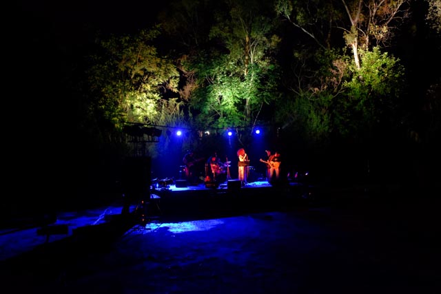 Η πολυμελής μπάντα Βερτζί Μαβί στη σκηνή του Our Festival με κιθάρα, ντραμς, μπουζούκια, μπαγλαμά και άλλα. Από πάνω τους τα δέντρα φωτισμένα αγκαλιάζουν τη σκηνή.