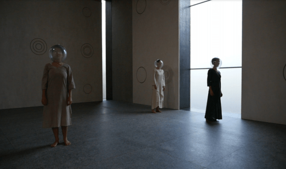 Σε ένα ψηλοτάβανο μεγάλο χώρο με ομόκεντρους κύκλους ζωγραφισμένους σε αραιή διάταξη στους τοίχους δεξιά και πίσω, τρεις γυναίκες με φορέματα μακριά στέκονται όρθιες κοιτώντας ευθεία μπροστά με μία διάφανη γυάλα τοποθετημένη στο κεφάλι τους κλειστή μέχρι το λαιμό.