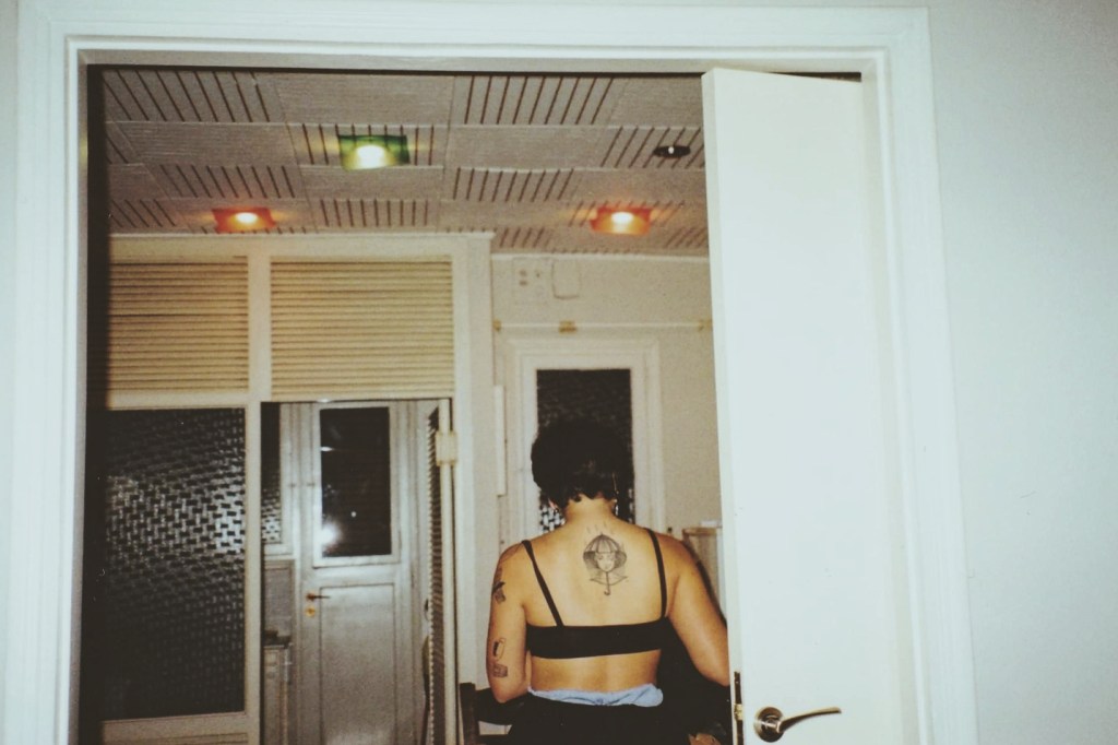 Από την ανοιγμένη πόρτα ενός δωματίου, φαίνεται η πλάτη ενός νέου κοριτσιού με κοντά μαλλιά. Φορά μαύρο μπουστάκι που αφήνει να φανούν τα τατουάζ στα μπράτσα και στο κέντρο της πλάτης της. Πρόκειται για το σχέδιο μιας κοπέλας που προστατεύεται από την βροχή μέσα από ένα καπέλο σε σχήμα ομπρέλας. 