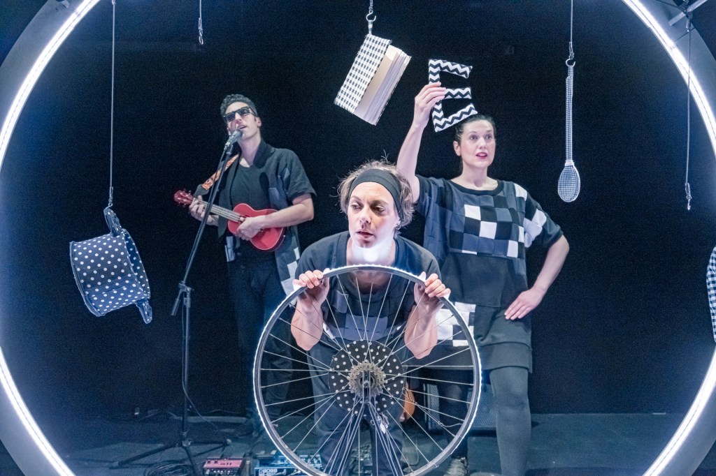 Σε μία μαύρη σκηνή, το περίγραμμα ενός μεγάλου κύκλου φωτίζεται από λευκό φως. Από μέσα του, ξεπροβάλλουν τρεις άνθρωποι. Δεξιά και πίσω, ένας άντρας με μαύρα γυαλιά παίζει γιουκαλίλι και τραγουδά σε ένα μικρόφωνο. Στο κέντρο και μπροστά μία γυναίκα με κορδέλα στα ξανθά κοντά της μαλλιά, κρατά με τα δυο της χέρια μια ρόδα ποδηλάτου και κοιτάζει προς τα αριστερά. Πίσω δεξιά, μια μελαχρινή γυναίκα με κοτσίδα κρατά ψηλά με το δεξί της χέρι ένα γράμμα Ε. όλοι τους φορούν μαύρα ρούχα με τετραγωνάκια στις αποχρώσεις του μαύρου, άσπρου και γκρι.